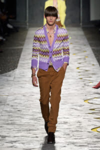 Male model walks the runway wearing a lavender zigzag cardigan over a purple shirt and brown trousers, with dark boots and jewelry.