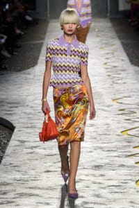 Model walking a runway in a lavender zigzag-knit polo and a glossy orange swirl-print skirt, carrying an orange handbag.