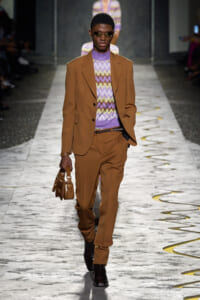 Male model on runway wearing a tan/brown suit with a purple zigzag knit top and brown shoes, carrying a small brown bag.