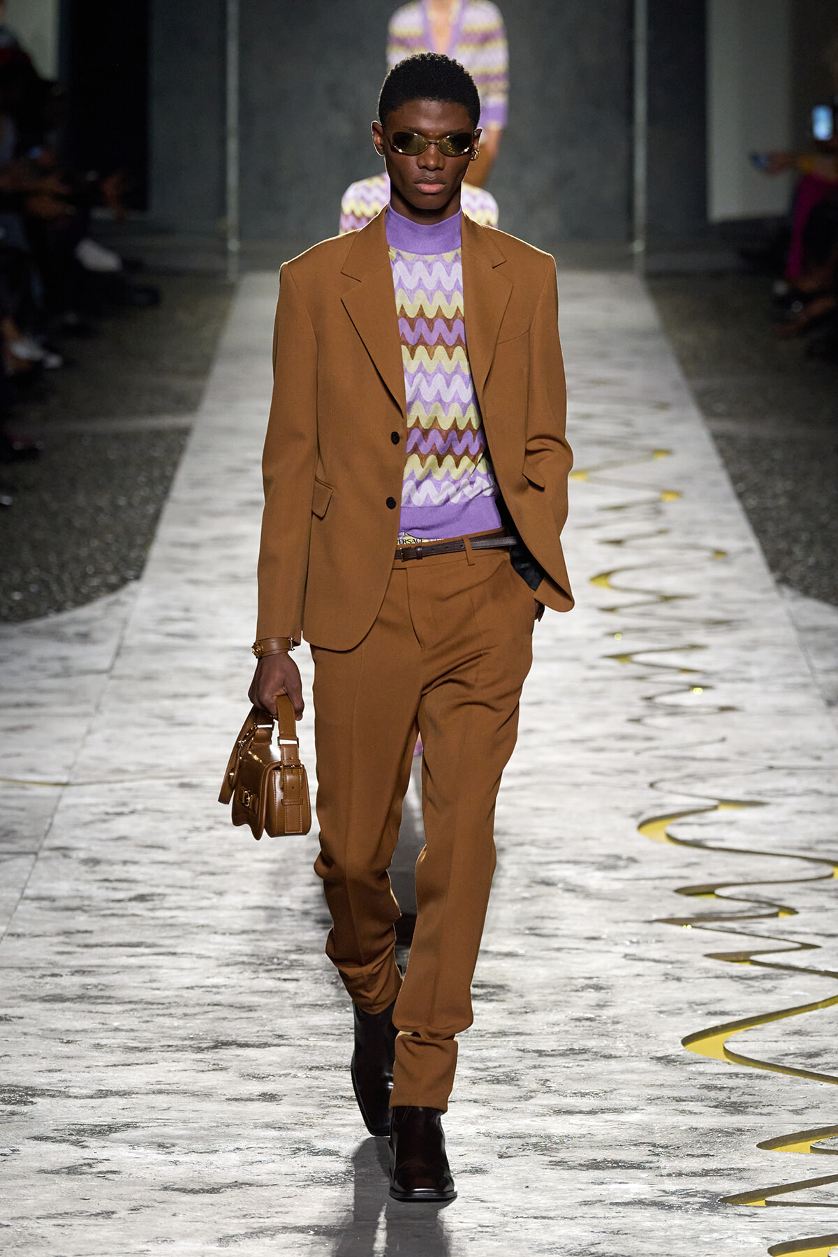 Male model on runway wearing a tan/brown suit with a purple zigzag knit top and brown shoes, carrying a small brown bag.