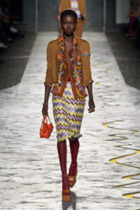 Model walking a fashion show runway in a mustard cardigan over a colorful patterned blouse and zigzag skirt, paired with burgundy tights, orange platform sandals, and a small orange handbag.