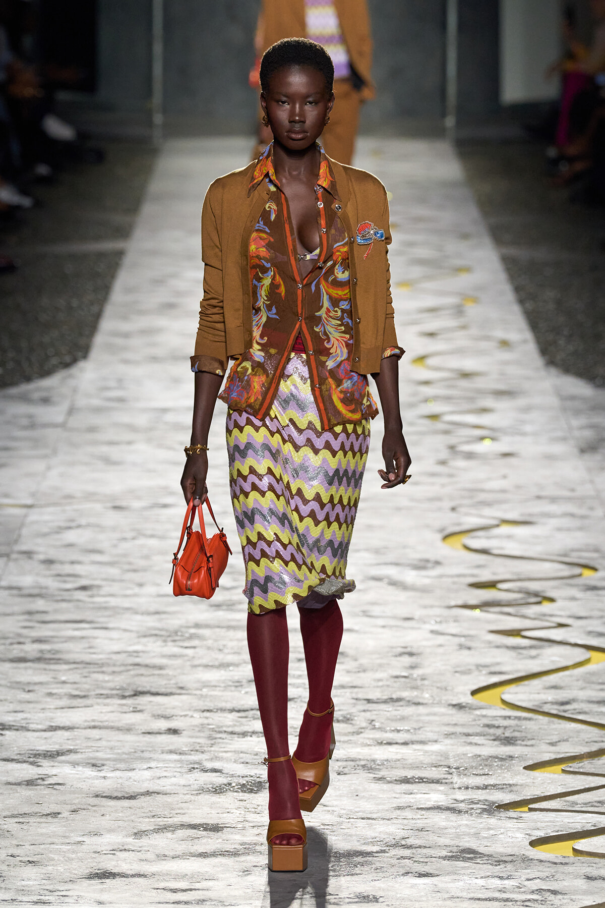 Model walking a fashion show runway in a mustard cardigan over a colorful patterned blouse and zigzag skirt, paired with burgundy tights, orange platform sandals, and a small orange handbag.