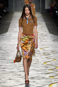 Model walks the runway in a brown polo with blue trim and a vibrant satin swirl skirt, carrying a brown handbag.
