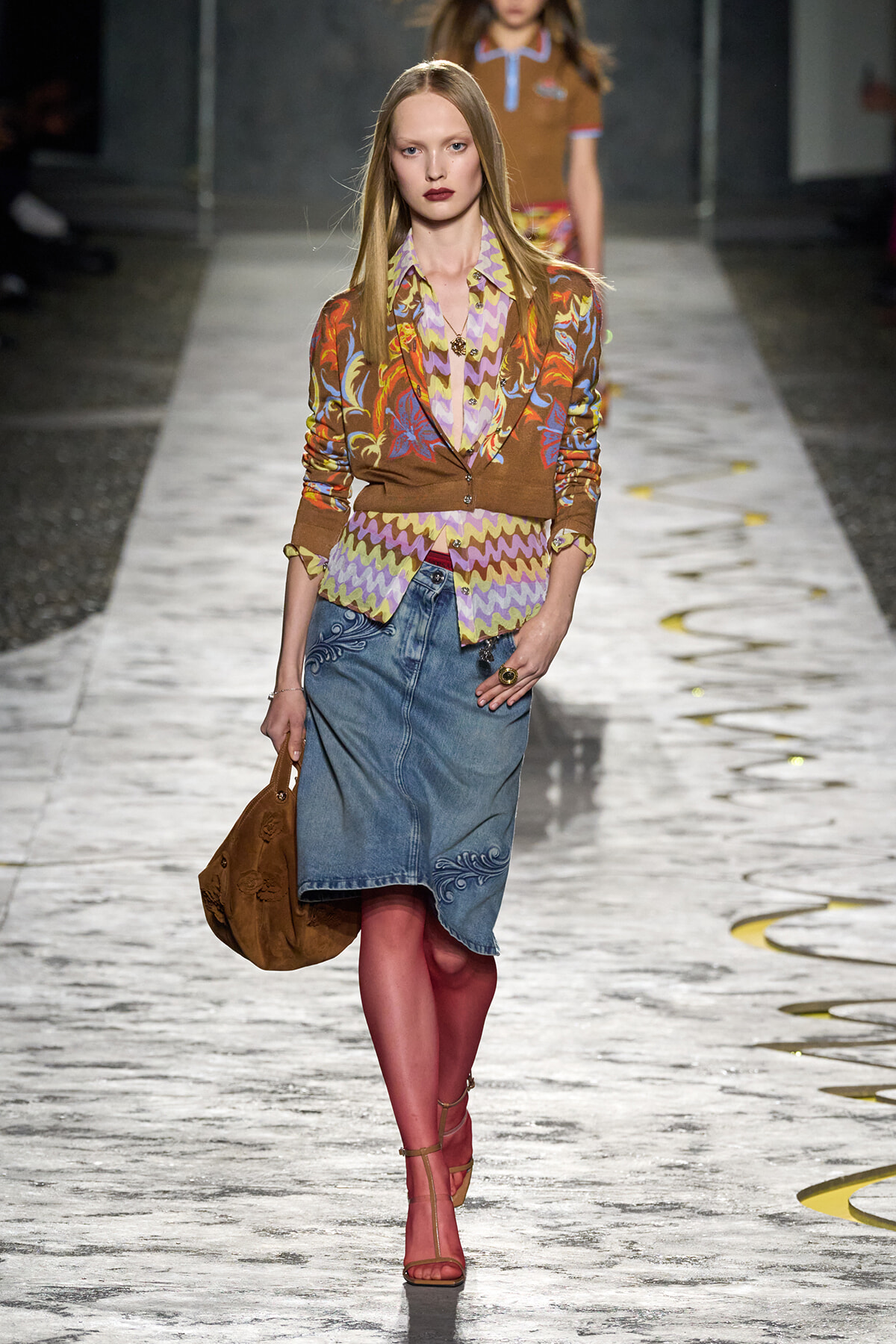 Model walks the runway in a brown floral cardigan over a pastel zigzag blouse, paired with a denim knee-length skirt and pink tights, carrying a brown suede bag.