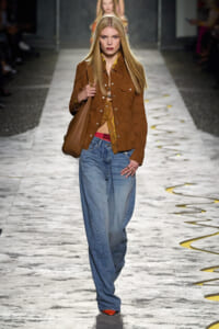 Model with long blonde hair walks a runway in a tan suede jacket, yellow top, and wide-leg jeans, carrying a brown shoulder bag.