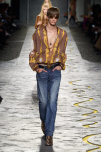Male model walks the runway in a brown paisley-print shirt, blue jeans, snakeskin boots, and sunglasses, hands in pockets.