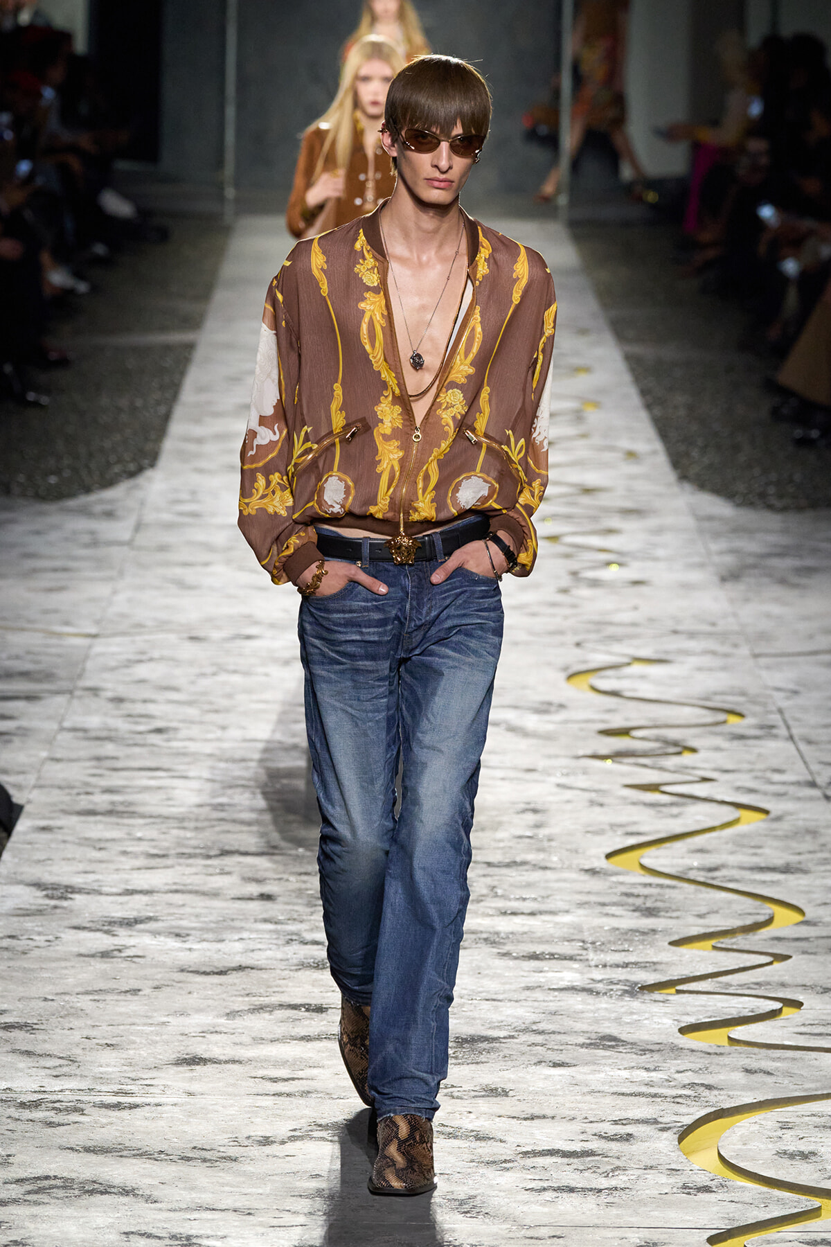 Male model walks the runway in a brown paisley-print shirt, blue jeans, snakeskin boots, and sunglasses, hands in pockets.