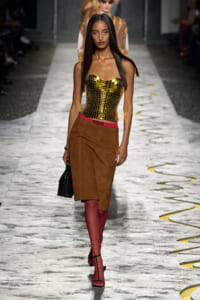 Model walks the runway in a gold strapless corset, brown suede skirt, red tights, and black handbag.