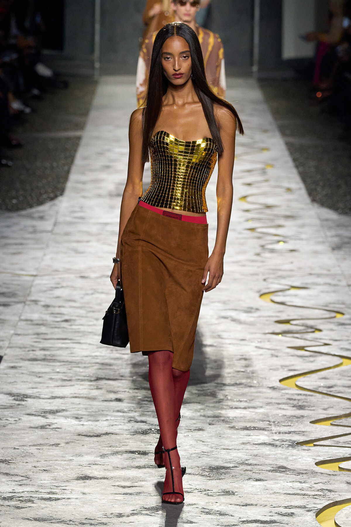 Model walks the runway in a gold strapless corset, brown suede skirt, red tights, and black handbag.