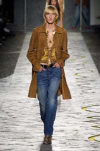 Male model walks a fashion runway in a brown suede trench over a floral-embroidered brown vest, blue jeans, and snakeskin boots.