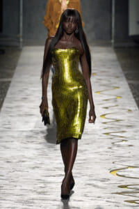 Dark-skinned model walks a runway in a strapless, gold mosaic dress with a fitted silhouette, holding a small clutch.