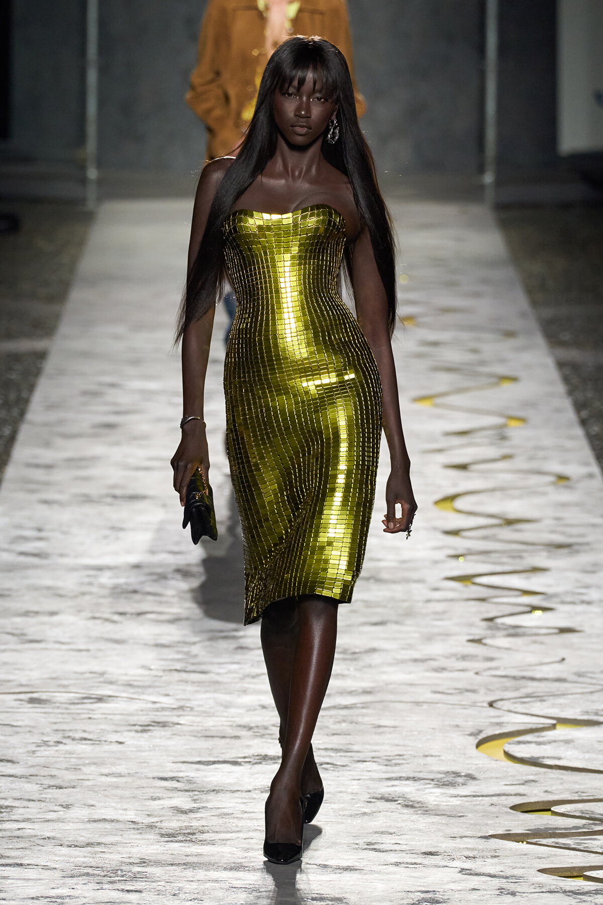 Dark-skinned model walks a runway in a strapless, gold mosaic dress with a fitted silhouette, holding a small clutch.