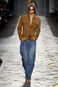 Model on a runway wearing a brown suede jacket, yellow patterned shirt, oversized blue jeans, brown boots, and sunglasses.