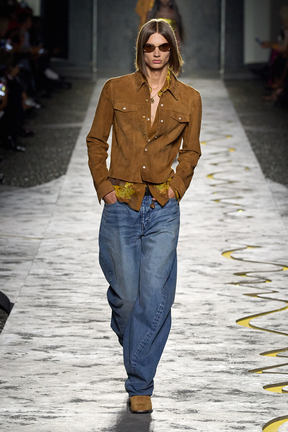 Model on a runway wearing a brown suede jacket, yellow patterned shirt, oversized blue jeans, brown boots, and sunglasses.
