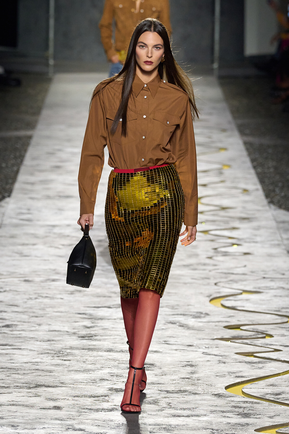 Model walking a fashion show runway in a brown button-down shirt with a metallic gold grid skirt, red belt, black handbag, and red strappy heels.