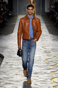 Male model walking a fashion runway in a brown leather jacket, blue shirt, jeans, and tan boots, carrying a small black bag