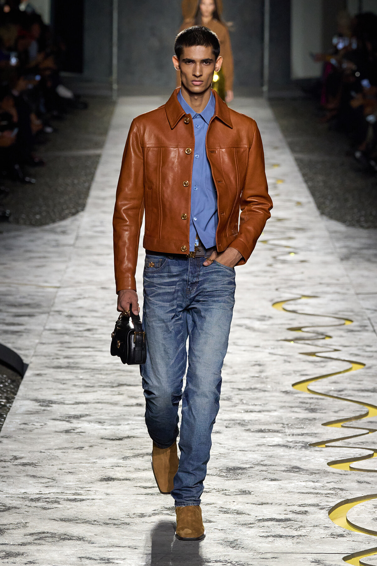 Male model walking a fashion runway in a brown leather jacket, blue shirt, jeans, and tan boots, carrying a small black bag