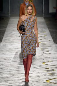 Model walking a runway in a one-shoulder brown dress with blue floral print, carrying a black quilted clutch and wearing strappy sandals.