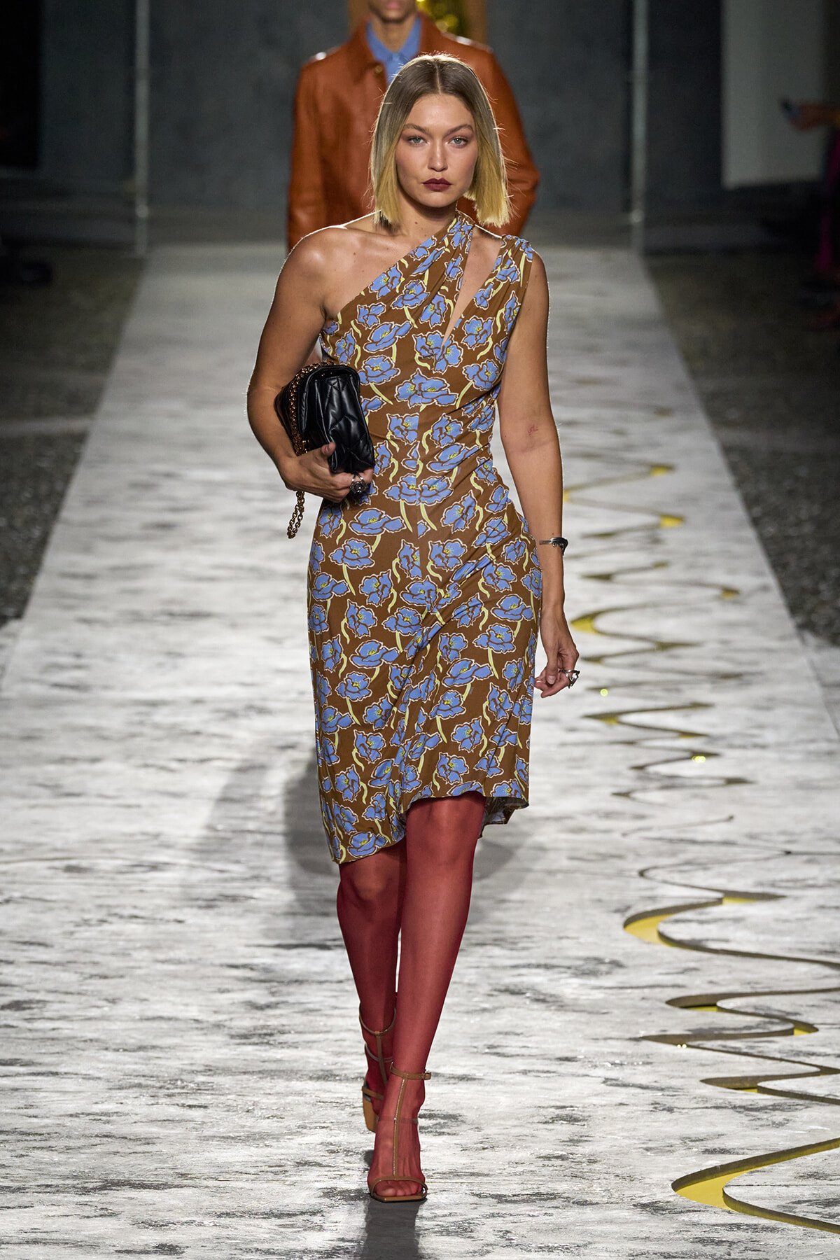 Model walking a runway in a one-shoulder brown dress with blue floral print, carrying a black quilted clutch and wearing strappy sandals.