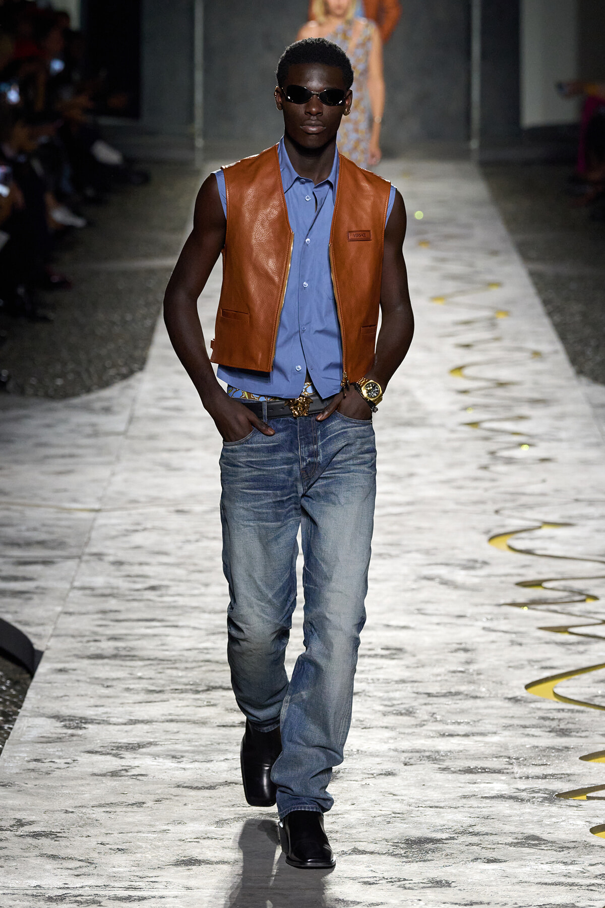 Male model wearing a tan leather vest over a blue button-down shirt on a runway, paired with washed jeans and black boots, sunglasses on.