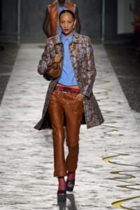 Model walking a runway in a brown floral coat over a blue button‑down shirt, brown leather pants, and platform sandals while carrying a brown bag.