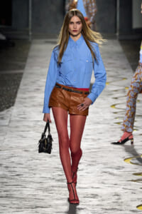 Runway model in a light blue button-down shirt and brown leather shorts with a red belt, carrying a small black handbag on a fashion catwalk.