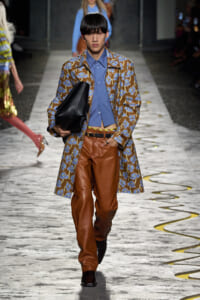 Male model walking a runway in a blue floral-brown coat, blue shirt, brown leather pants, and a black portfolio clutch.