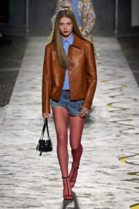 Model walks the runway in a brown leather jacket over a blue shirt, denim shorts, pink tights, black strappy heels, and a small black handbag.