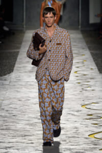 Male model on a fashion runway wearing a brown and blue floral suit, holding a dark brown bag over his chest.