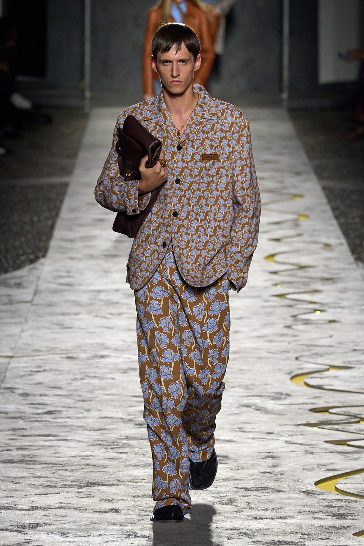 Male model on a fashion runway wearing a brown and blue floral suit, holding a dark brown bag over his chest.