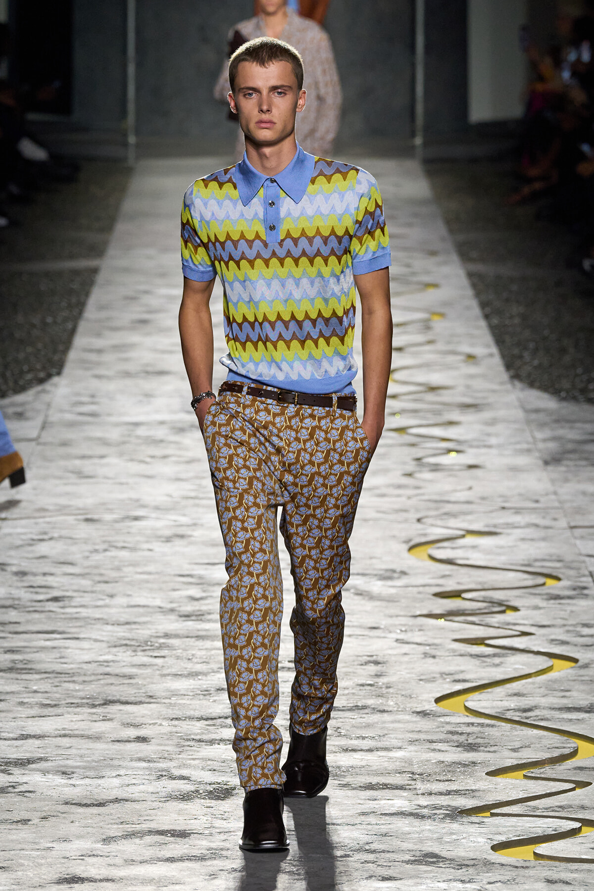 Male model walking a fashion runway in a blue, multicolored zigzag polo shirt and tan patterned trousers with brown belt.