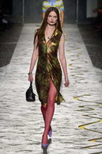 Model walking the runway in a metallic camouflage-pattern sleeveless dress with a front slit, paired with lilac heels and a small black handbag.