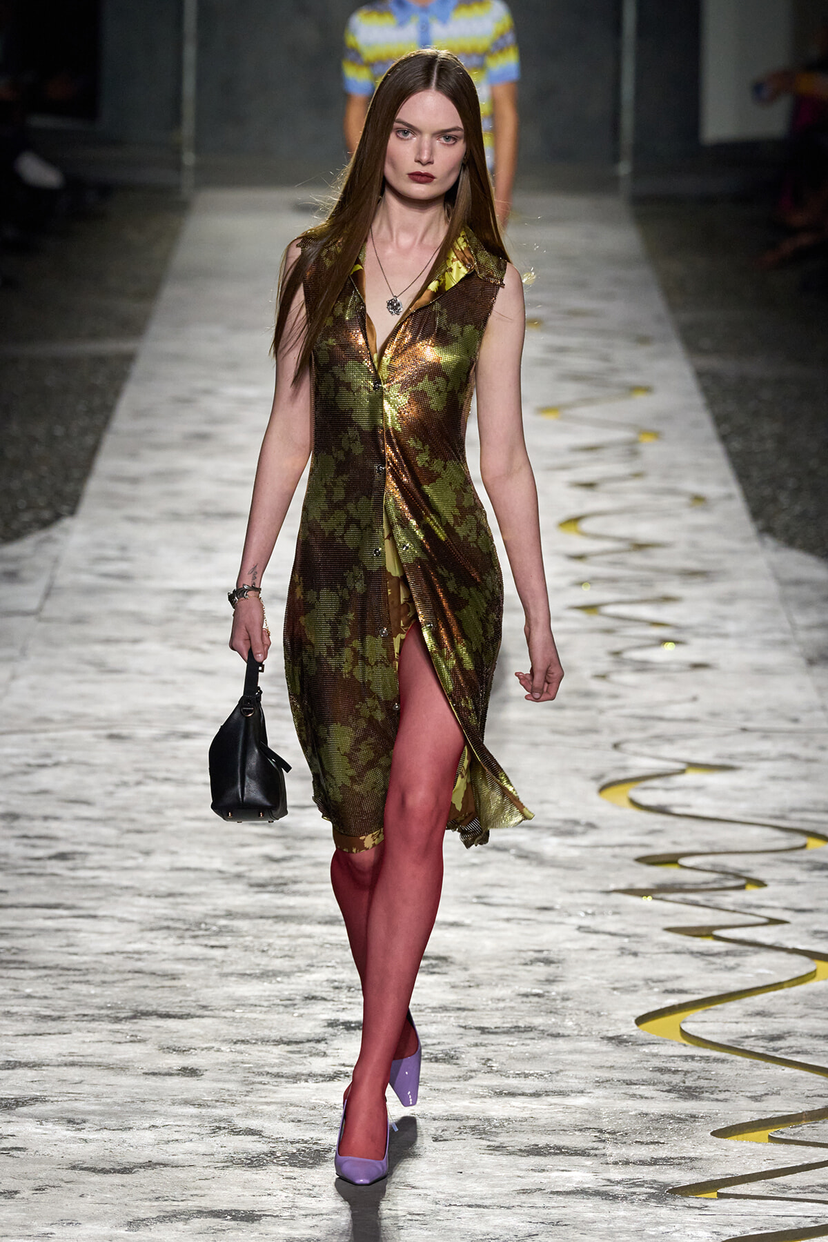 Model walking the runway in a metallic camouflage-pattern sleeveless dress with a front slit, paired with lilac heels and a small black handbag.