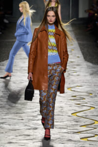 Model walking a fashion runway in a caramel brown leather coat over a blue zigzag-knit top, paired with blue floral trousers and a small black handbag.