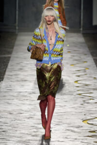 Model on a runway wearing a blue, yellow, and purple zigzag cardigan over a metallic camouflage skirt, tan heels, and a brown quilted handbag.