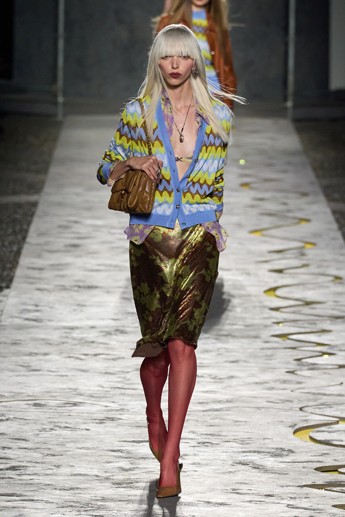 Model on a runway wearing a blue, yellow, and purple zigzag cardigan over a metallic camouflage skirt, tan heels, and a brown quilted handbag.