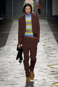 Model on a fashion runway wearing a brown suit over a multicolor zigzag knit sweater, with tan boots and a black bag in hand.