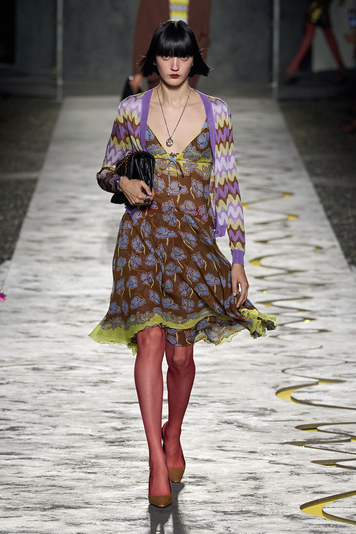 Model walking a runway in a brown floral dress with a lime-green hem, layered with a purple zigzag cardigan, and carrying a black clutch.