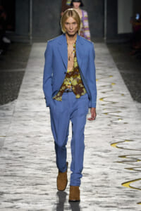 Model walks the runway in a light blue suit with a yellow-brown floral shirt and brown suede boots.