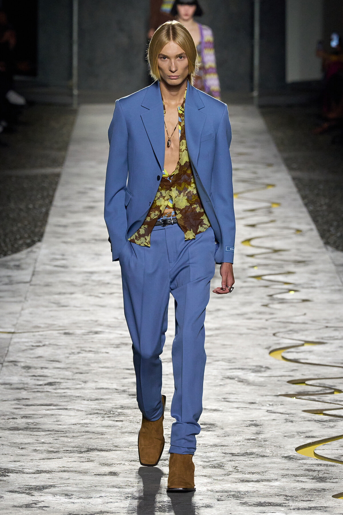 Model walks the runway in a light blue suit with a yellow-brown floral shirt and brown suede boots.