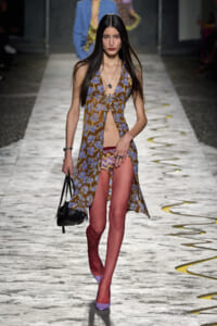 Model walks a fashion runway in a brown-blue floral halter dress with a front slit, lavender heels, and a black handbag.