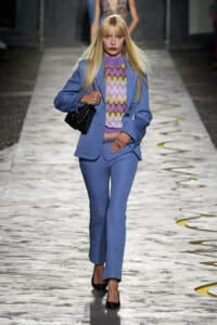 Model walking a fashion runway in a powder-blue pantsuit with a lavender zigzag knit top and a black quilted handbag.