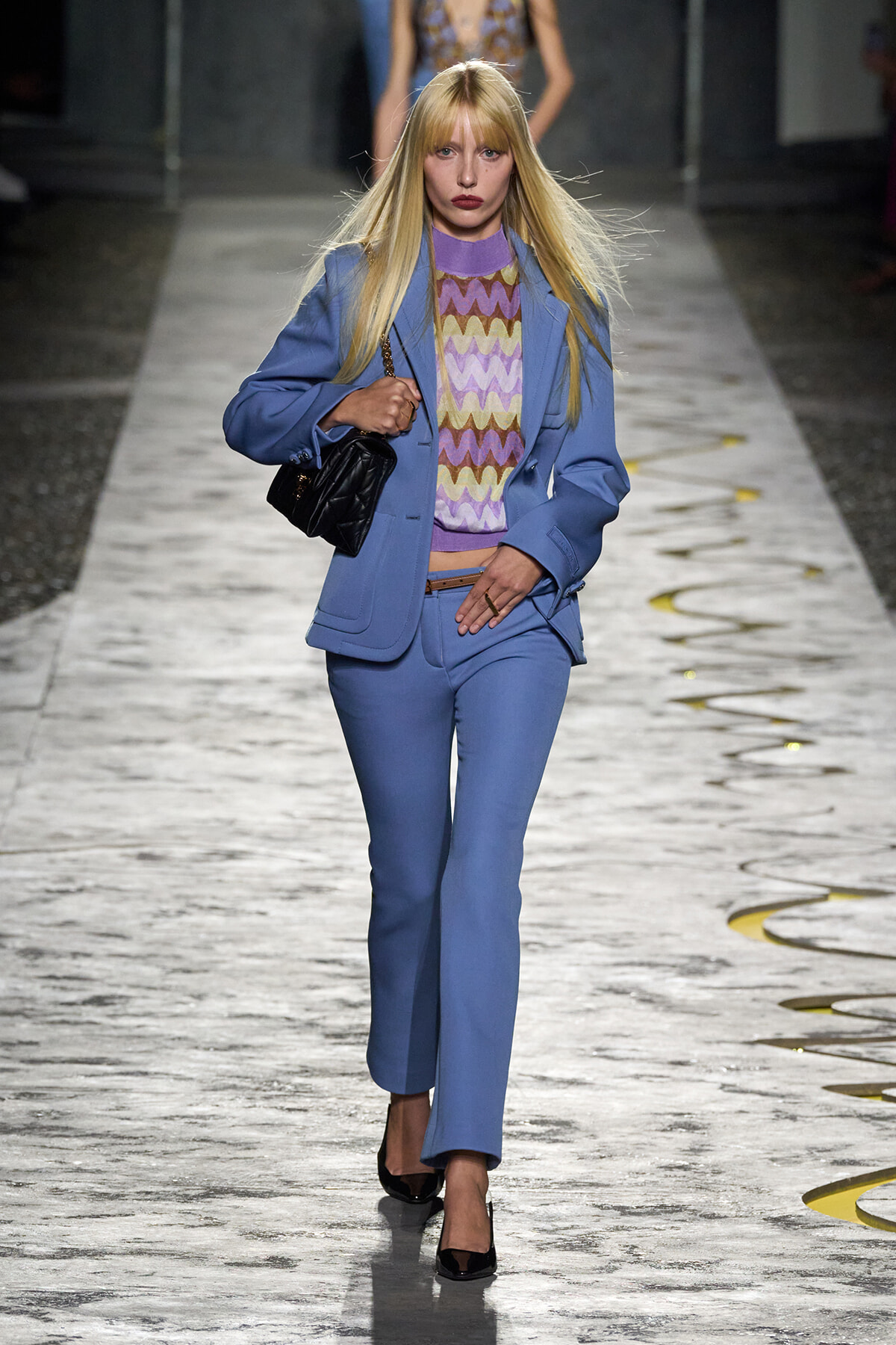 Model walking a fashion runway in a powder-blue pantsuit with a lavender zigzag knit top and a black quilted handbag.