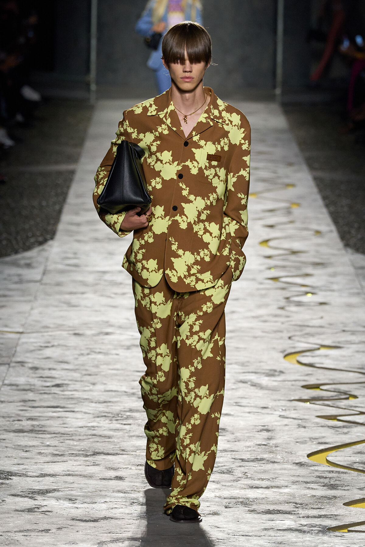 Runway model in a brown suit with pale lime floral print, carrying a black handbag on the catwalk.