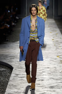 Male fashion model on the runway wearing a light blue overcoat over a bold multicolored patterned shirt, with brown trousers and tan suede boots, holding a small dark clutch.