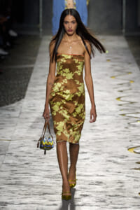 Model walking runway in a brown satin strapless dress with yellow floral pattern and yellow heels, carrying a small colorful handbag.