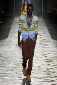 Male model walks the runway wearing a blue, yellow, and brown zigzag cardigan with a deep neckline, paired with maroon trousers and tan boots, accessorized with a pendant necklace.