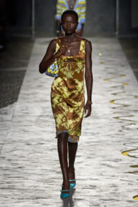 Model walking runway in a brown-yellow floral midi dress with tie-front detail, turquoise heels, and a small patterned handbag.