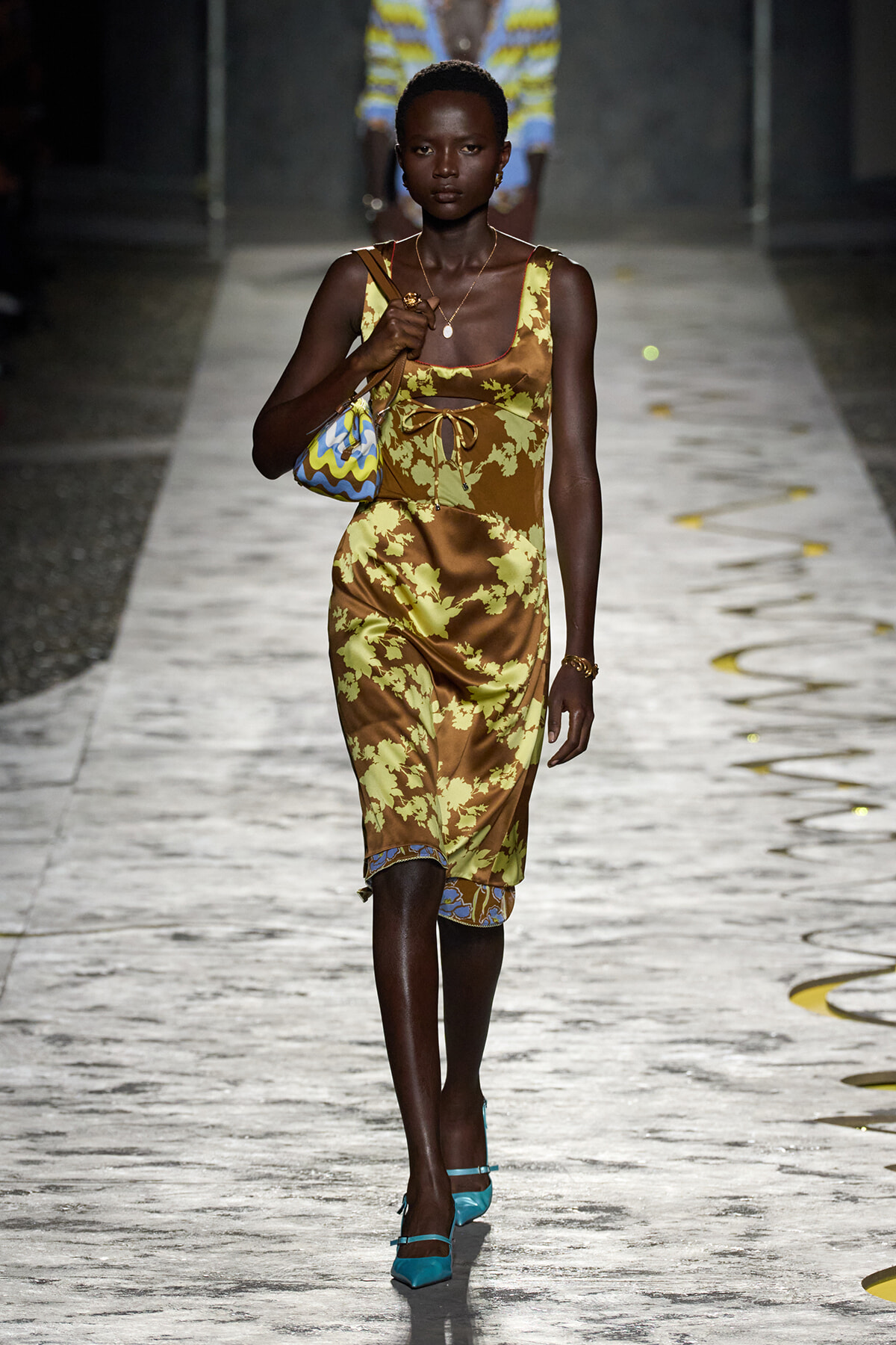 Model walking runway in a brown-yellow floral midi dress with tie-front detail, turquoise heels, and a small patterned handbag.