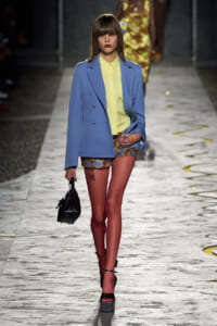 Model walking the runway in a light blue oversized blazer over a pale yellow shirt, paired with floral shorts, red tights, black platform sandals, and a small black handbag.